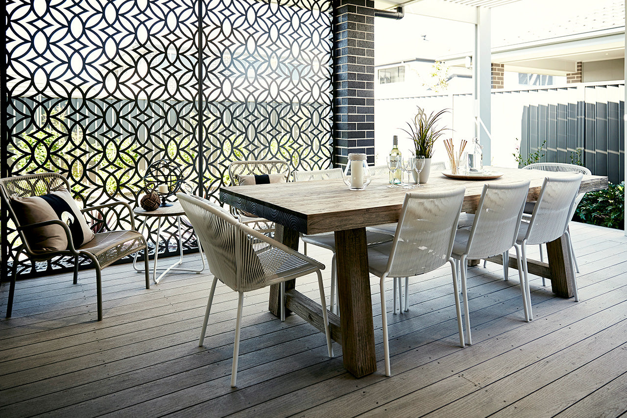 Powder-coated steel garden privacy screen with laser-cut decorative design, set in a stylish patio as a backdrop to an outdoor table and chairs, adding privacy and modern backyard décor.